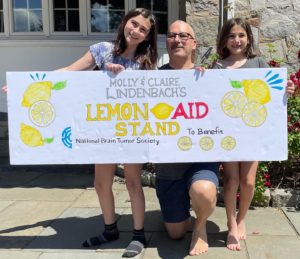 An adult male holds up a Lemon-Aid Stand banner alongside his two 7 and 11-year-old daughters.