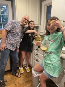 An adult male puts a lemon in his mouth to make a silly smile alongside his 7 and 11-year-old daughters.